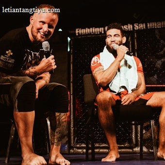 Two individuals are seated on a stage, each holding a microphone and smiling. They are in a sports-related setting with one wearing a casual black outfit and the other in sportswear, with a towel around his neck. The background includes elements of a fighting or martial arts cage.
