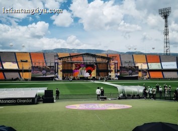 A sports stadium with a large stage set up on the field, hosting an outdoor event. Spectators wearing colorful hats sit in the foreground. The seating areas display vibrant colors, and a clear blue sky with scattered clouds is visible. Large screens display graphics related to the event.