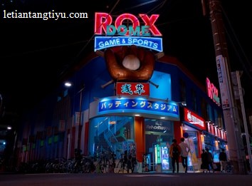 A vibrant, neon-lit facade of a gaming and sports establishment at night, featuring a large glove holding a baseball above the entrance. The building is adorned with colorful signs in both English and Japanese, with various advertisements and an illuminated sign that says 'ROX DOME'. Several people are walking towards the entrance, and bicycles are parked along the side.